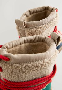 Flauschige beige Stoffstiefel mit roten Schnürsenkeln, strukturiertem Kragen und Nähdetails. Das Innenfutter scheint weich und gemütlich zu sein.