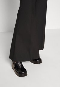 Svarta vida byxor som täcker skarpa svarta plattformsskor på en ljusgrå golv och en enkel bakgrund.