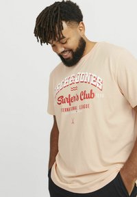 Světle broskvové bavlněné tričko s červeným grafickým textem "Surfer's Club" a "International League." Krátké rukávy a volný střih.