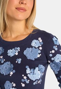 Marineblauer Pullover mit gerippter Textur, der geschichtete Blumenapplikationen in hellblauen und weißen Farbtönen aufweist, asymmetrisch auf der Vorderseite angeordnet.
