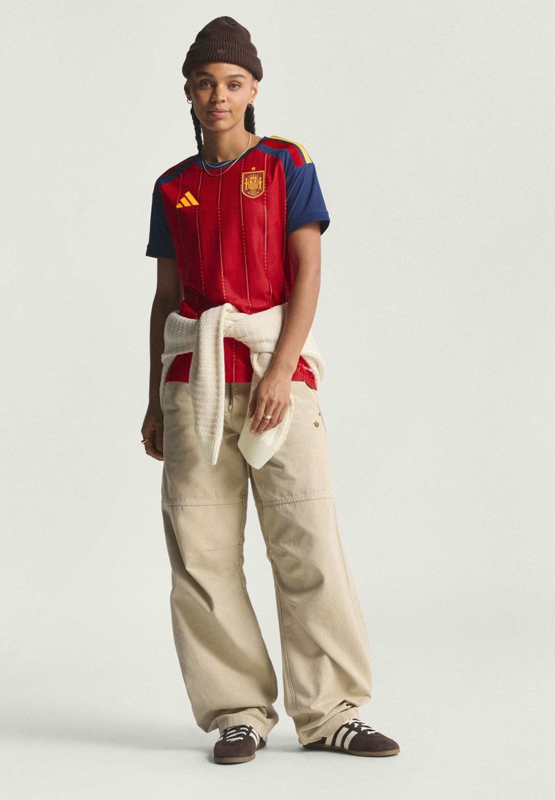 Rood en blauw voetbalshirt met korte mouwen en verticale strepen, beige wijde broek, en bruine sneakers met witte strepen, afgewerkt met een bruine muts.