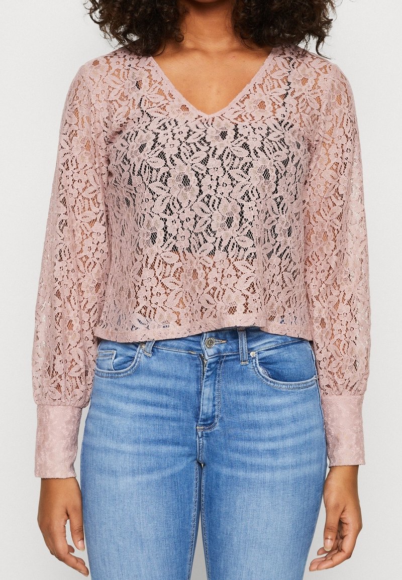 Femme portant un chemisier en dentelle florale rose clair à manches longues avec un col en V et des manches retroussées, associé à un jean bleu clair.