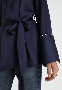 Top de satén navy con un lazo en la cintura, mangas anchas y costuras blancas en los puños. Combinado con jeans de mezclilla.