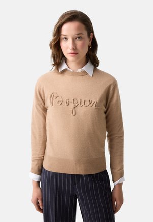 Vrouw draagt een beige trui met verhoogde tekst "Pourquoi", een witte blouse met kraag eronder en marineblauwe krijtstreepbroek op een witte achtergrond.