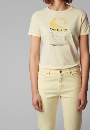 Femme portant un t-shirt graphique jaune pâle à manches courtes avec l'inscription « Summer Love » et un jean jaune pâle assorti sur un fond gris.