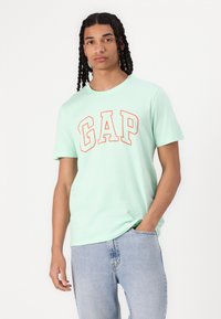 Camiseta verde menta de algodón, con letras "GAP" en naranja vibrante. Mangas cortas y cuello redondo. Llevada con jeans de color azul claro.