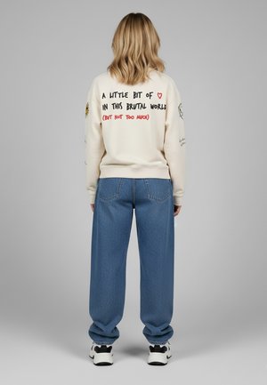 Felpa crema con testo grafico sulla schiena, abbinata a jeans blu larghi e scarpe da ginnastica bianche. Presenta dettagli ricamati sulle maniche.