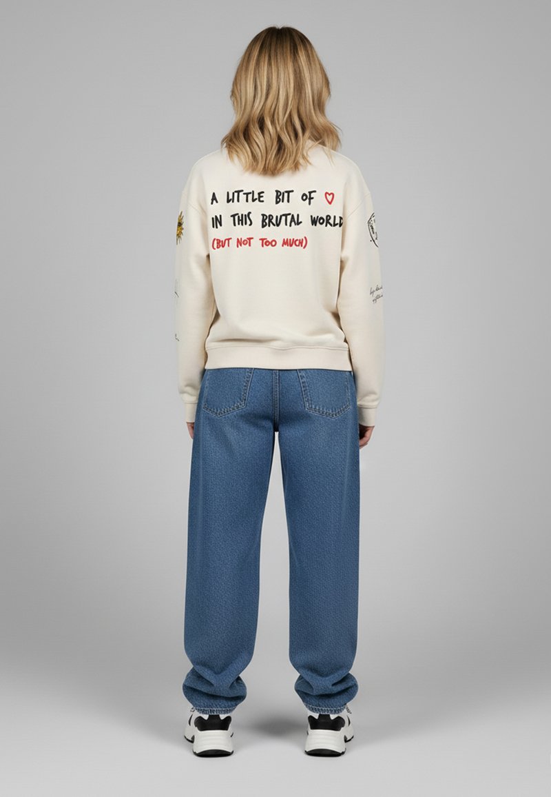 Felpa crema con testo grafico sulla schiena, abbinata a jeans blu larghi e scarpe da ginnastica bianche. Presenta dettagli ricamati sulle maniche.