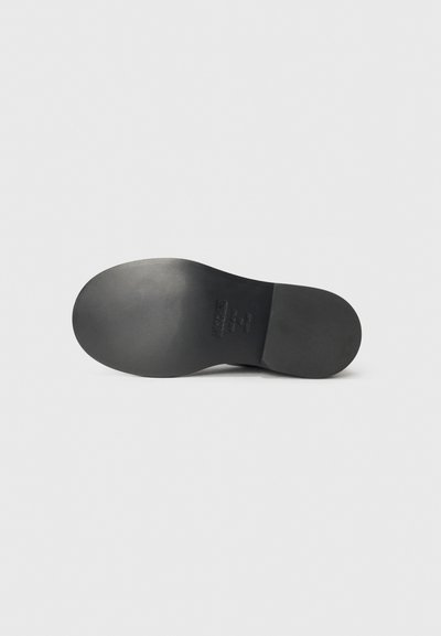 Suela de zapato negra con una superficie lisa, punta ligeramente redondeada y detalles de marca en relieve cerca del talón. Diseño minimalista, sin patrones.