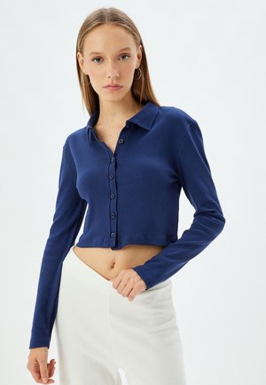 Camisa corta de color azul marino con botones y mangas largas, textura acanalada y un cuello clásico. Cuenta con seis botones a juego en la parte frontal.