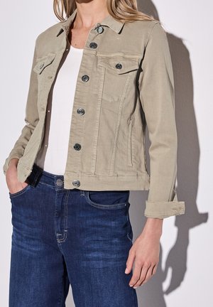 Vrouw met een beige jasje met knopen over een wit shirt en donkerblauwe spijkerbroek, met één hand in de zak en een ontspannen pose.