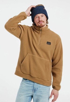 Brauner Fleece-Pullover mit Reißverschlusskragen, Fronttasche und kleinem schwarzen Logo. Getragen mit hellblauen Jeans und einer dunkelblauen Rippmütze.