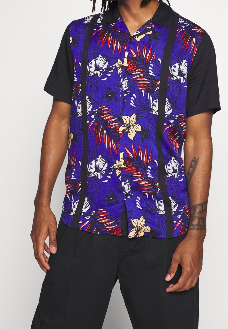 Homme portant une chemise à manches courtes boutonnée avec des manches noires, un devant à motif floral bleu et un pantalon noir.