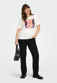 Valkoinen T-paita, jossa on värikäs graafinen printti, joka esittää naisen kasvoja; yhdistetty mustiin suoriin housuihin ja teksturoituun mustaan käsilaukkuun.