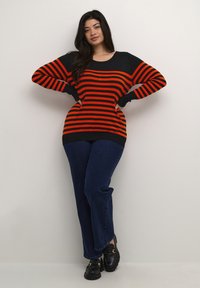 Zwart en rood gestreepte sweater met lange mouwen, wijde halslijn en een ruime pasvorm, gecombineerd met donkere spijkerbroek en zwarte instappers.