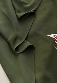 Donkergroene stof met zachte plooien en een zichtbare rand, met een gedeeltelijk bordeauxrood en wit bladpatroon aan de rechteronderkant.