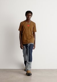 Brun t-skjorte med en rød etikett, paret med fargeblokkerte jeans med blå og svarte lapper, og brune slipper-sko; gående posering.