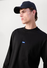 Zwarte lange mouwen shirt met een ronde hals en een klein blauw "HUGO" logo op de borst, gecombineerd met een bijpassende zwarte baseballpet.
