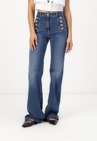 Ψηλόμεσα φαρδιά τζιν παντελόνια σε σκούρο τζιν με έξι χρυσά κουμπιά σε κάθε πλευρά και ομαλή υφή. Εφαρμοστό κάτω από τα γόνατα.