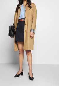 Trench-coat beige avec boutons en bois, jupe bleu marine foncé, blouse bleu clair, sac à main noir et chaussures à talons noires pointues avec une finition texturée.