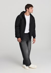 Giacca nera con cappuccio e chiusura con zip, indossata sopra una maglietta bianca, abbinata a jeans scuri e sneakers bianche. Design semplice, senza motivi visibili.