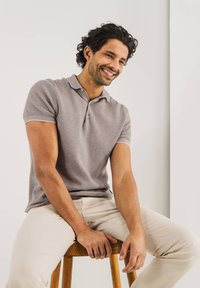 Polo shirt van lichtgrijze gebreide stof, met een klassieke kraag, korte mouwen met contrasterende afwerking. Model zittend op een houten kruk.