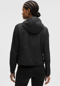 Sort zip-jakke med hætte, lavet af letvægts, tekstureret stof. Har elastiske manchetter og en indsnøret talje for et tætsiddende look.
