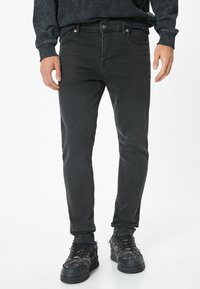 Jeans de mezclilla negra con corte slim, con una cintura de altura media, diseño de cinco bolsillos y una sutil textura desgastada. Combinados con zapatillas negras.