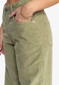 Olivengroene corduroy broek met een hoge taille, zijzakken en een subtiele ribbelstructuur. Een enkele knop en metalen hardwaredetail zichtbaar.