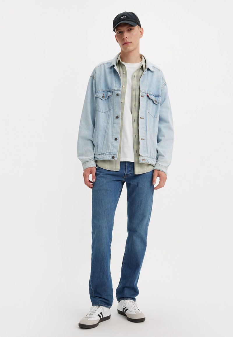 Helles blauer Denim-Jacke über einem grün karierten Hemd und einem weißen T-Shirt, kombiniert mit blauen Jeans und weißen Sneakers mit schwarzen Akzenten.