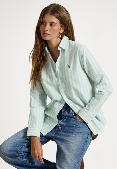 Gaiši zaļa svītraina pogājama krekla ar baltām un rozā detaļām, ar klasisku apkakli un garām piedurknēm, kopā ar zilām džinsām.