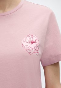 Ανοιχτό ροζ κοντομάνικο μπλουζάκι με λεπτό σκουρόροζ περίγραμμα λουλουδιού στο στήθος, που φοράει ένα άτομο.