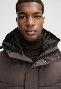 Giacca imbottita marrone con colletto in pelliccia sintetica, dotata di chiusura con zip e dettagli in bottone. Indossata sopra un dolcevita marrone scuro.