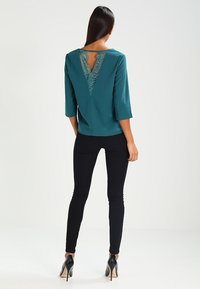 Femme de dos, portant un haut bleu sarcelle avec un détail en dentelle transparente en V dans le dos, un pantalon skinny noir et des chaussures à talons noirs sur fond blanc.