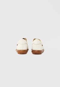 Par av vita läderskor i casual stil med bruna gummisulor, sedda bakifrån mot en enkel ljusgrå bakgrund.