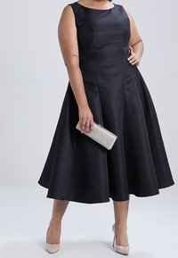 Svart ärmlös klänning med figurnära överdel och utsvängd kjol. Slät tyg med en subtil lyster. Matchas med en silvrig clutch och nudefärgade klackar.