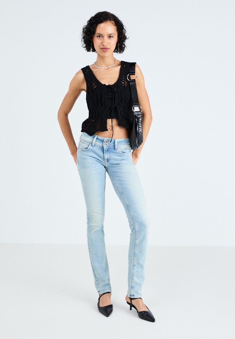 Top corto all'uncinetto nero con un nodo sul davanti, abbinato a jeans aderenti blu chiaro e mule nere a punta. Collana in argento.