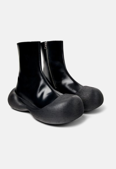 Svarta ankelstövlar med en glänsande ovandel, dragkedjestängning och en distinkt grov texturerad sula. Rund tådesign.