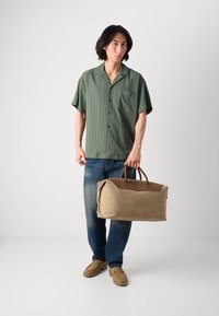 Green striped short-sleeve shirt, blue jeans, and beige suede duffel bag with brown leather accents. Model holds the bag with one hand.
