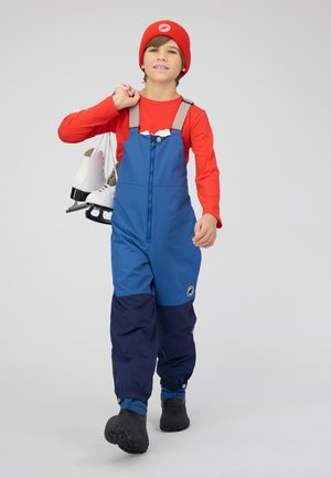 Blue and navy waterproof overall with adjustable straps, zipper front, and logo, paired with a red long-sleeve shirt and black boots.