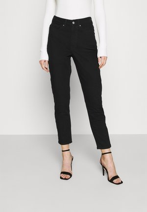 Vrouw draagt zwarte high waist skinny jeans, wit geribd langermig shirtje en zwarte hoge hak sandalen met bandjes, staand tegen een witte muur.