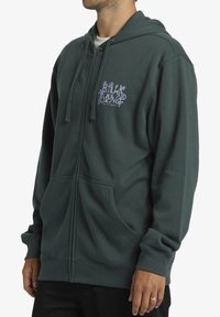 Sudadera verde con cremallera hecha de una suave mezcla de algodón, con un bolsillo frontal, capucha con cordón y un logotipo gráfico púrpura en el lado izquierdo del pecho.