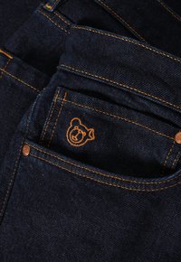 Jeans in denim blu scuro con cuciture arancioni e un logo di orso ricamato sulla tasca anteriore. Texture liscia e design classico.