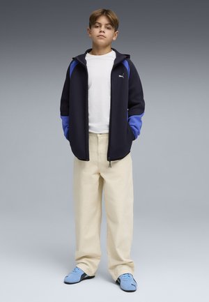 Giovane in piedi che indossa una maglietta bianca, una giacca con zip nera e blu, pantaloni beige ampi e sneakers azzurro chiaro su sfondo grigio.