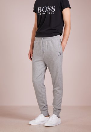 Mężczyzna w szarych spodniach dresowych ze ściągaczami przy kostkach, białych trampkach i czarnej koszulce z logo "BOSS HUGO BOSS".