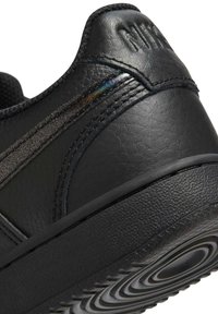 Close-up van de hak van een zwarte leren Nike-sneaker met een geëmboste logo en een rubberen zool met opvallend rond profiel.