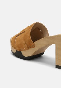 Bruine suède klomp met een houten hak en een zwarte rubberen zool. Heeft een uitgesneden ontwerp op de bovenkant en contrasterende stiksels op de randen.