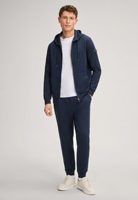 Navy Trainingsanzug-Set mit einer Kapuzenjacke zum Reißverschluss und passenden Jogginghosen. Hergestellt aus einem weichen Material, mit Taschen ausgestattet und kombiniert mit weißen Sneakers.