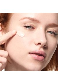 Femme rousse à la peau claire applique une crème sur la joue avec le doigt, illustrant l'application de soins de la peau et un maquillage naturel éclatant.