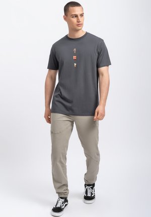 T-shirt gris à manches courtes avec un design graphique sur le devant, associé à un pantalon beige clair et des baskets noires. Texture du tissu lisse.